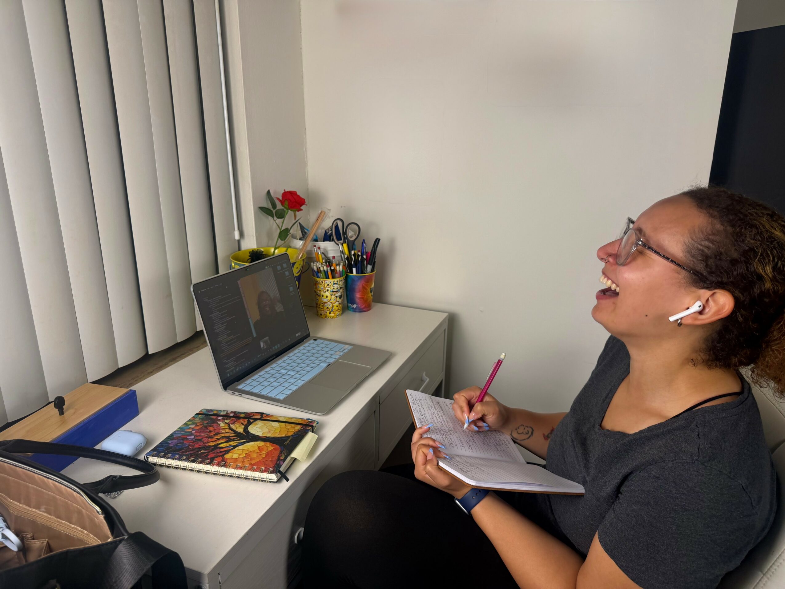 Woman sitting at desk, smiling, writing in notebook, with laptop and office supplies nearby.