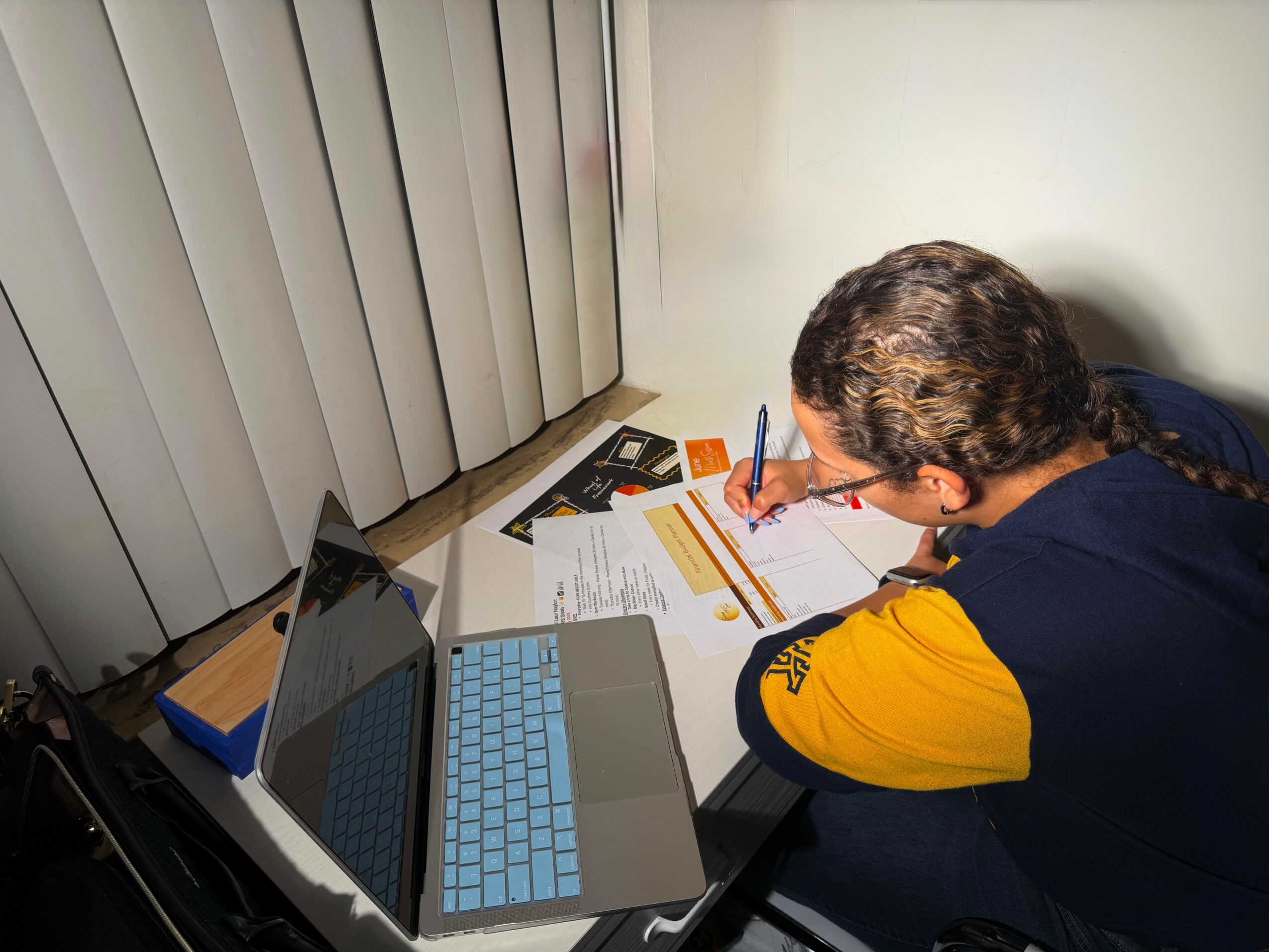Person with dark hair and yellow sleeve writing at desk with laptop, papers, and books, near vertical blinds.