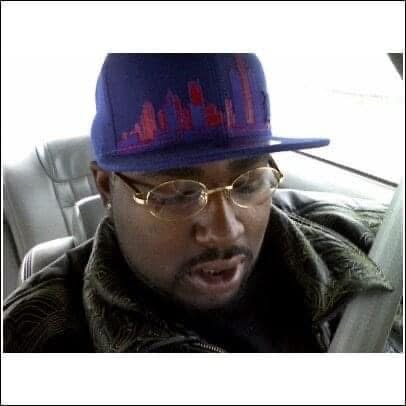 Person wearing a blue cap with a city skyline design, glasses, and a leather jacket, sitting in a vehicle.