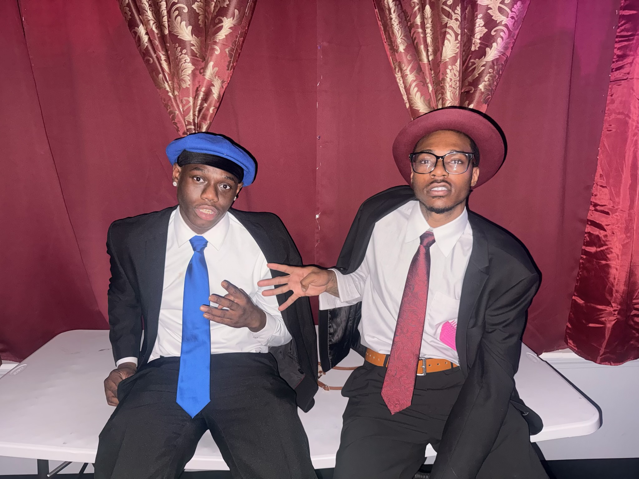 Two men in suits and hats sitting in front of a red curtain, one with a blue hat and the other with a pink hat, gesturing with hands.