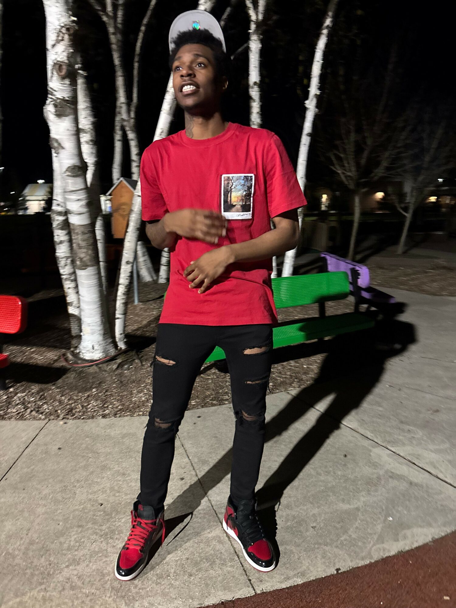 Person standing outdoors on sidewalk, wearing red shirt, black pants, and sneakers, with trees and a bench in background.