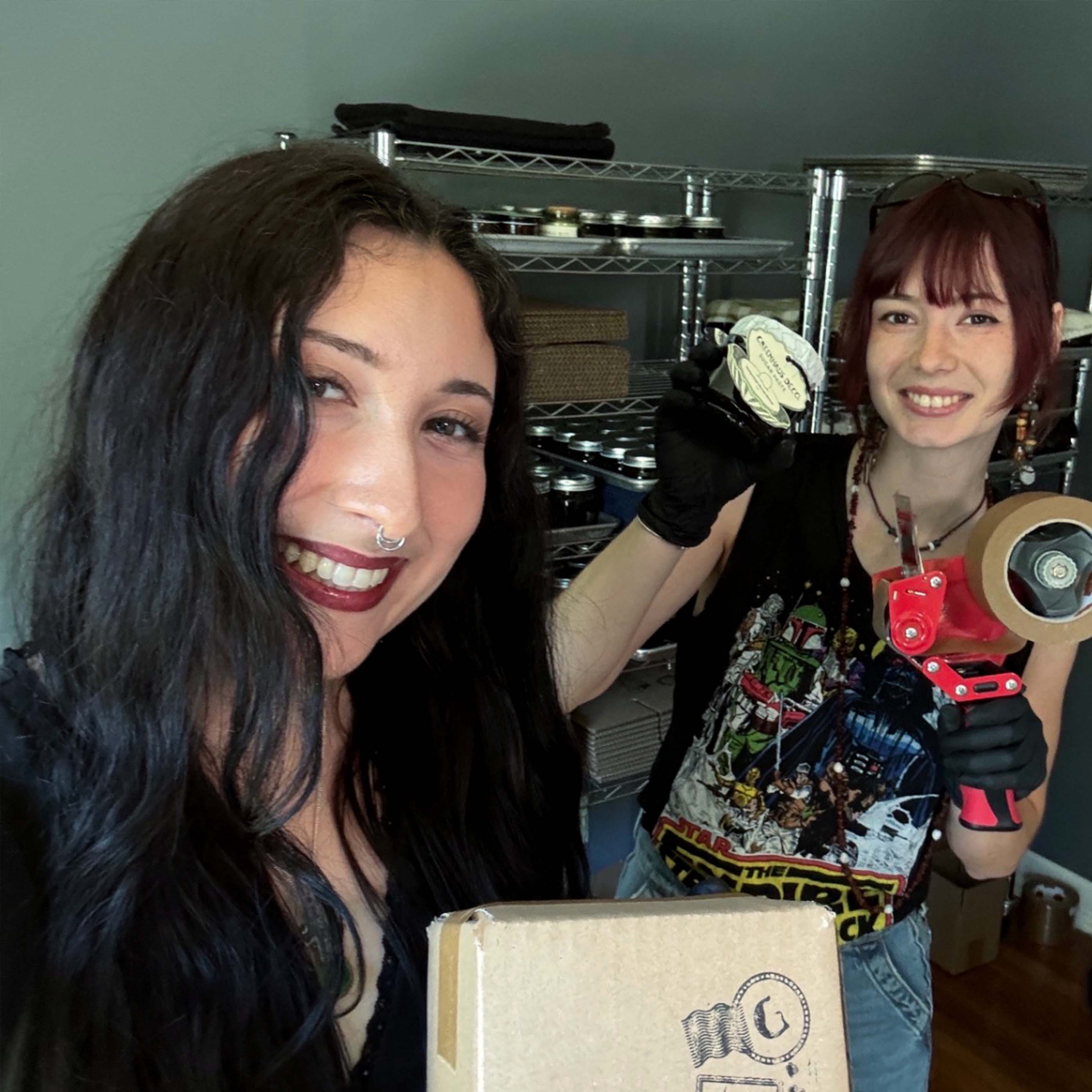 Two women smiling, one holding a cake and a camera, in a room with metal shelves.