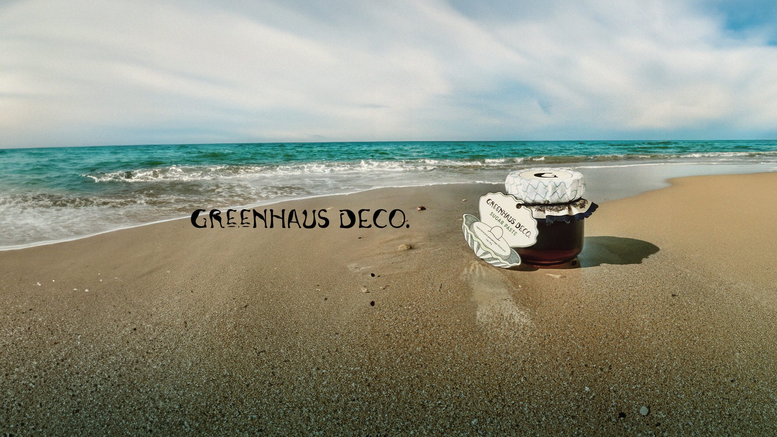 Beach scene with a painted sign on the sand reading 'GREENHAUS DECO' and a white object nearby, ocean and sky in background.