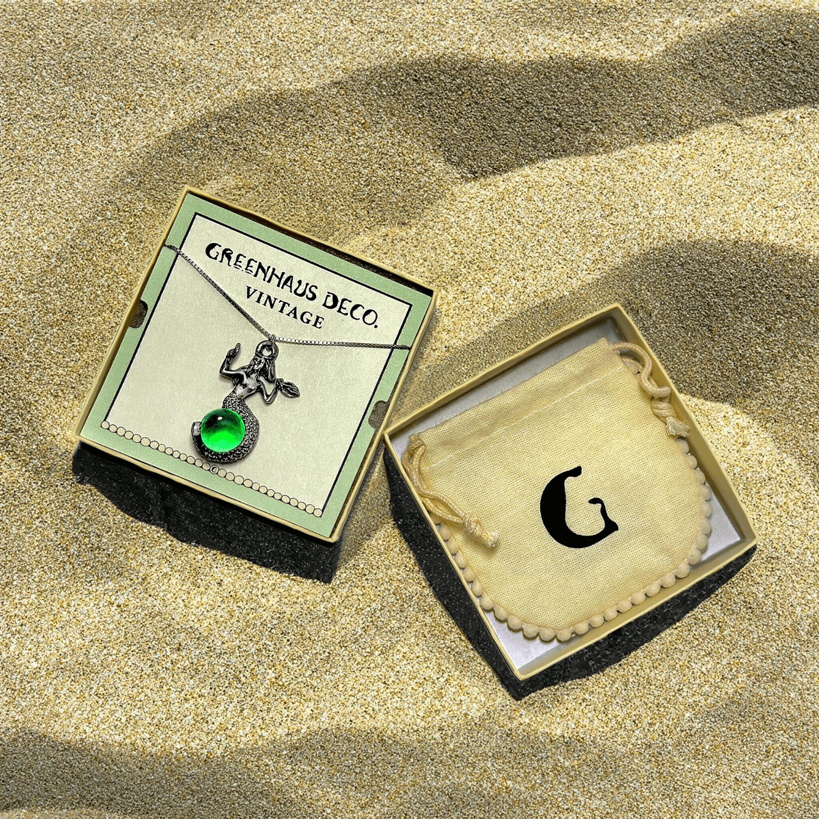 Two jewelry boxes on sand, one with a green pendant and a small figure, the other with a letter G cookie.