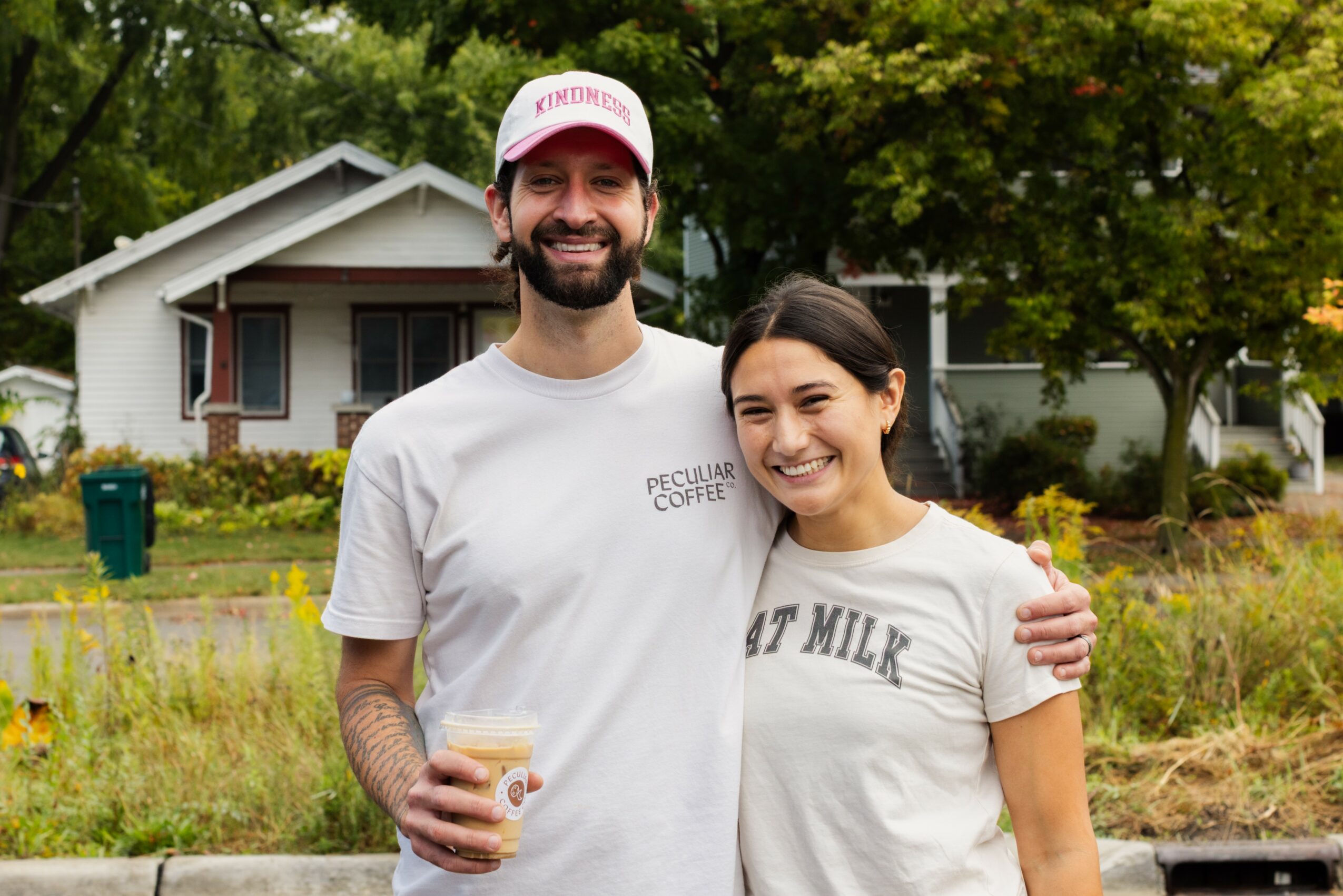 A smiling man and woman stand outdoors in front of a house, with trees and grass behind them.
