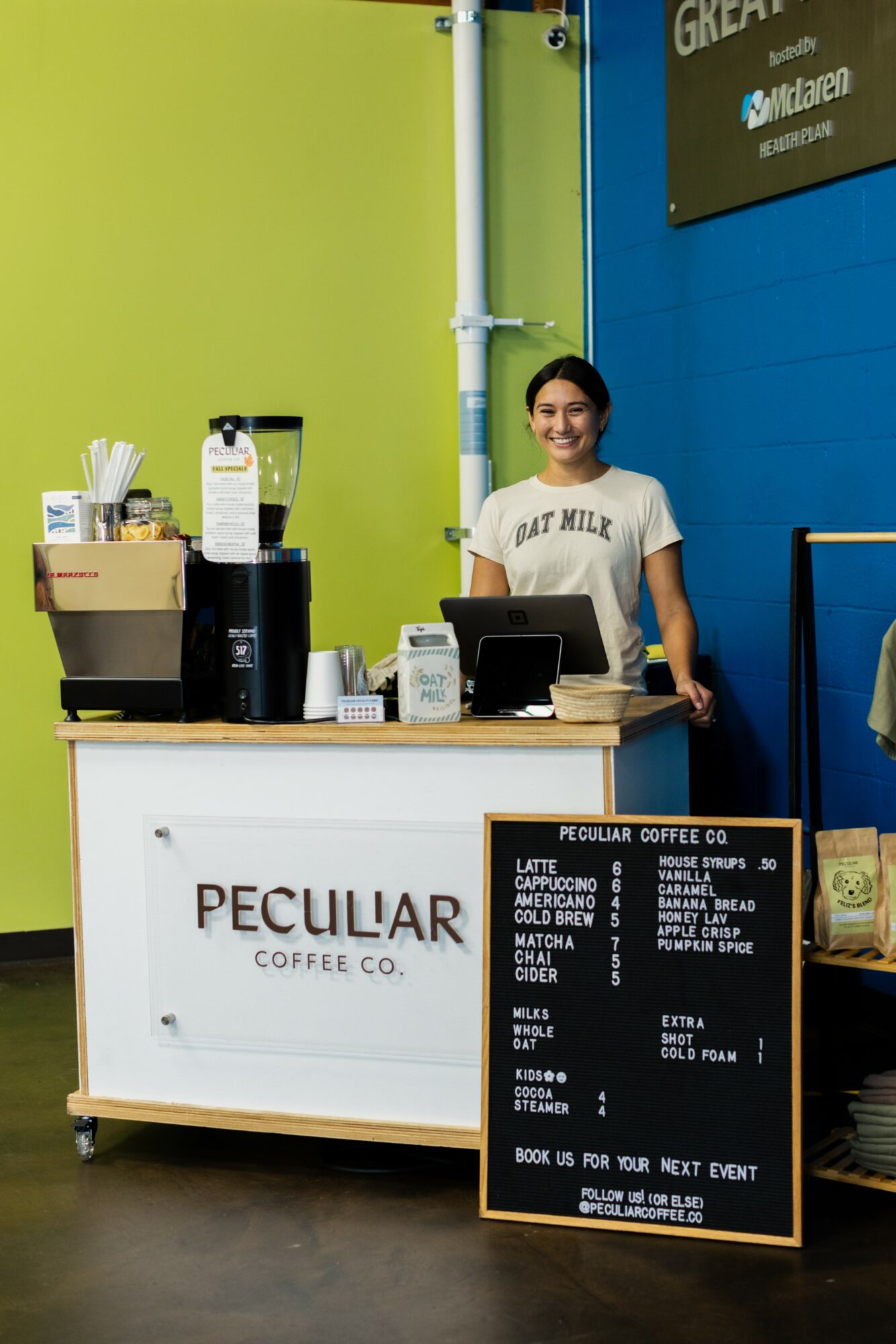 Person standing behind a coffee counter with a blackboard menu, in a brightly colored cafe, smiling.