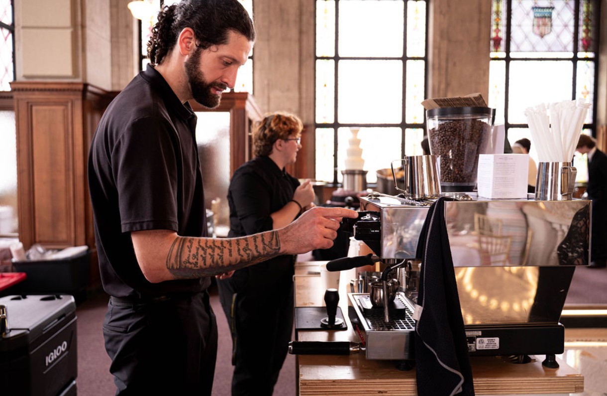 Barista with tattoos preparing coffee at a counter in a cafe, with a woman in the background, large windows, and wooden decor.
