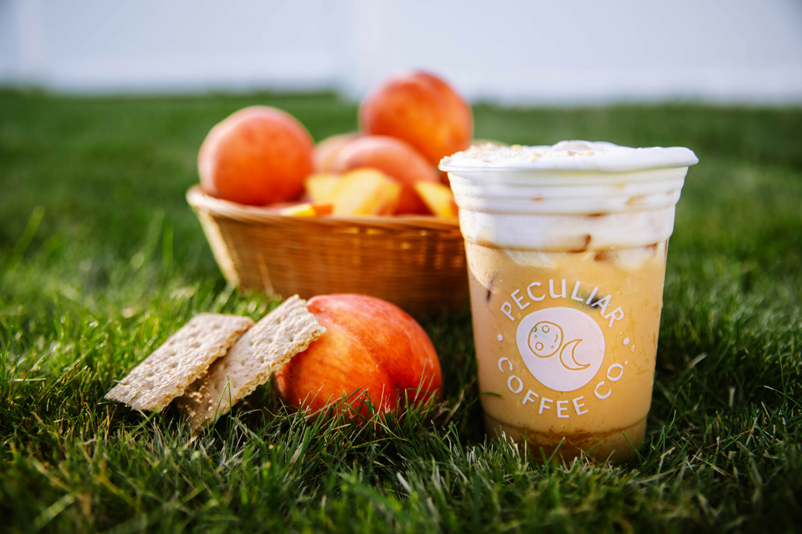 Basket of apples and peaches, two slices of bread, and a coffee drink on grass outdoors.