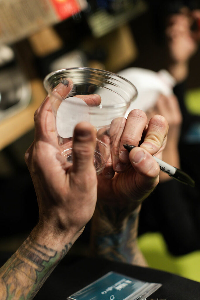 Person signing a clear glass object with a black marker, tattoos visible on arm, blurred background.