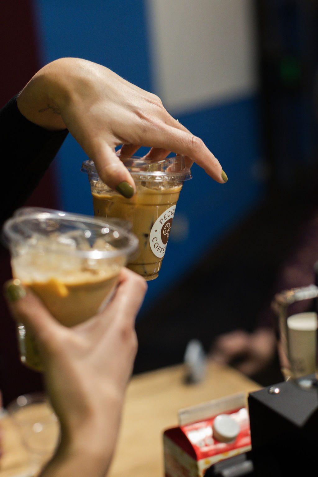 Person's hand holding a plastic cup with iced coffee, another hand reaching for the drink, background blurred.