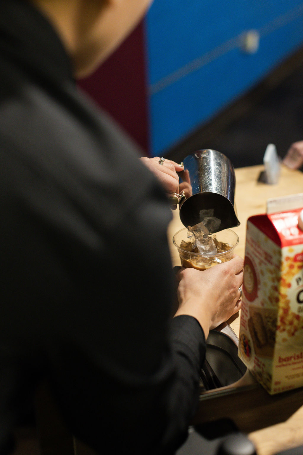 Person pouring a beverage from a metal pitcher into a glass on a wooden table, with a cereal box nearby.
