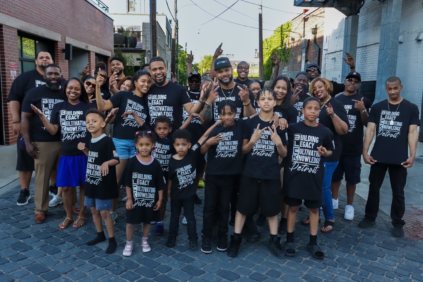 Group of people, including children and adults, standing on a street, smiling and wearing matching black T-shirts.