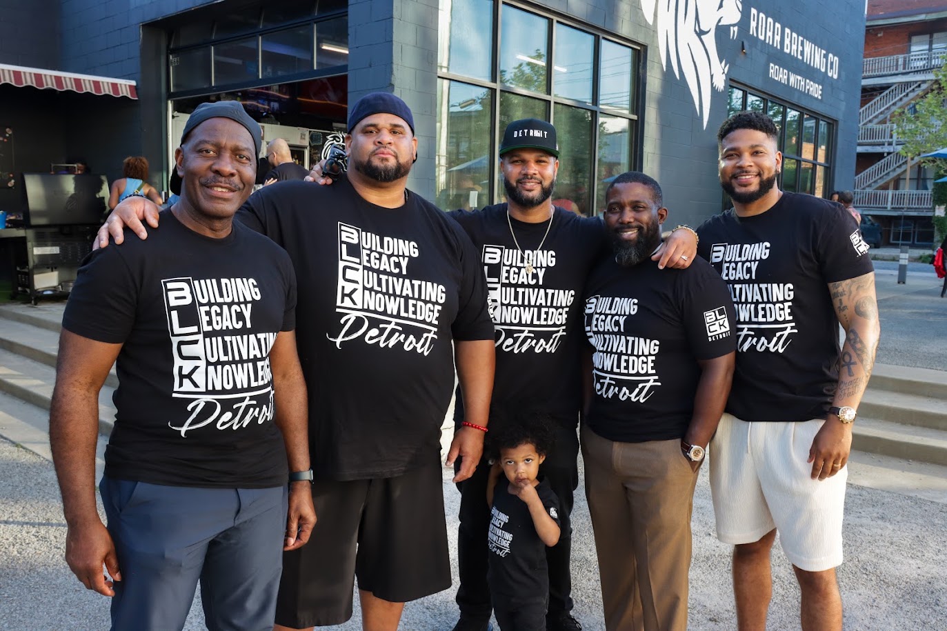Group of five men and one child standing outside a building, smiling, wearing matching black shirts with white text.