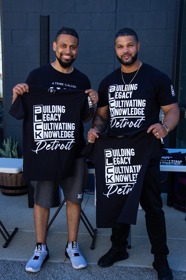 Two men standing outdoors holding black T-shirts with white text, smiling, with a dark wall background.
