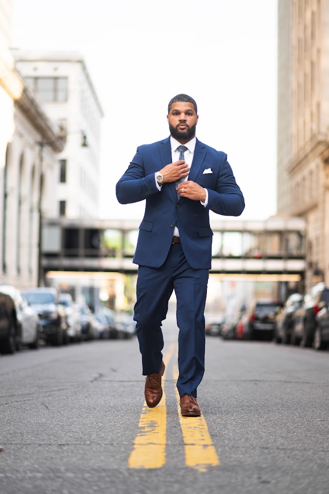 Man in a navy suit walking on a city street, adjusting his tie, with buildings and parked cars in the background.
