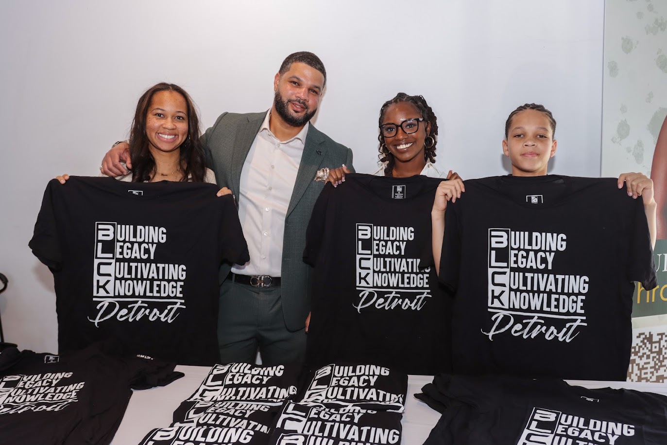 Four people smiling and holding black T-shirts with white text, standing behind a table with similar T-shirts, in a bright room.