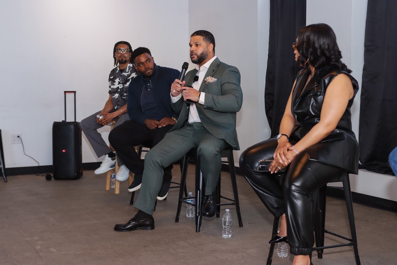 Four people seated on chairs in a panel discussion, with one person speaking into a microphone, in a room with black curtains.