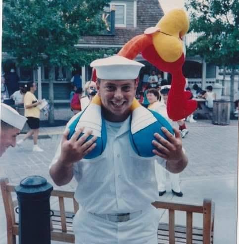 Person in sailor costume holding plush duck hat, smiling, outdoor setting with people and trees in background.