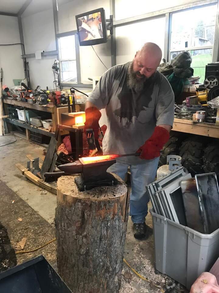 Man working with glowing metal on an anvil in a workshop, with tools and equipment around, inside a well-lit space.