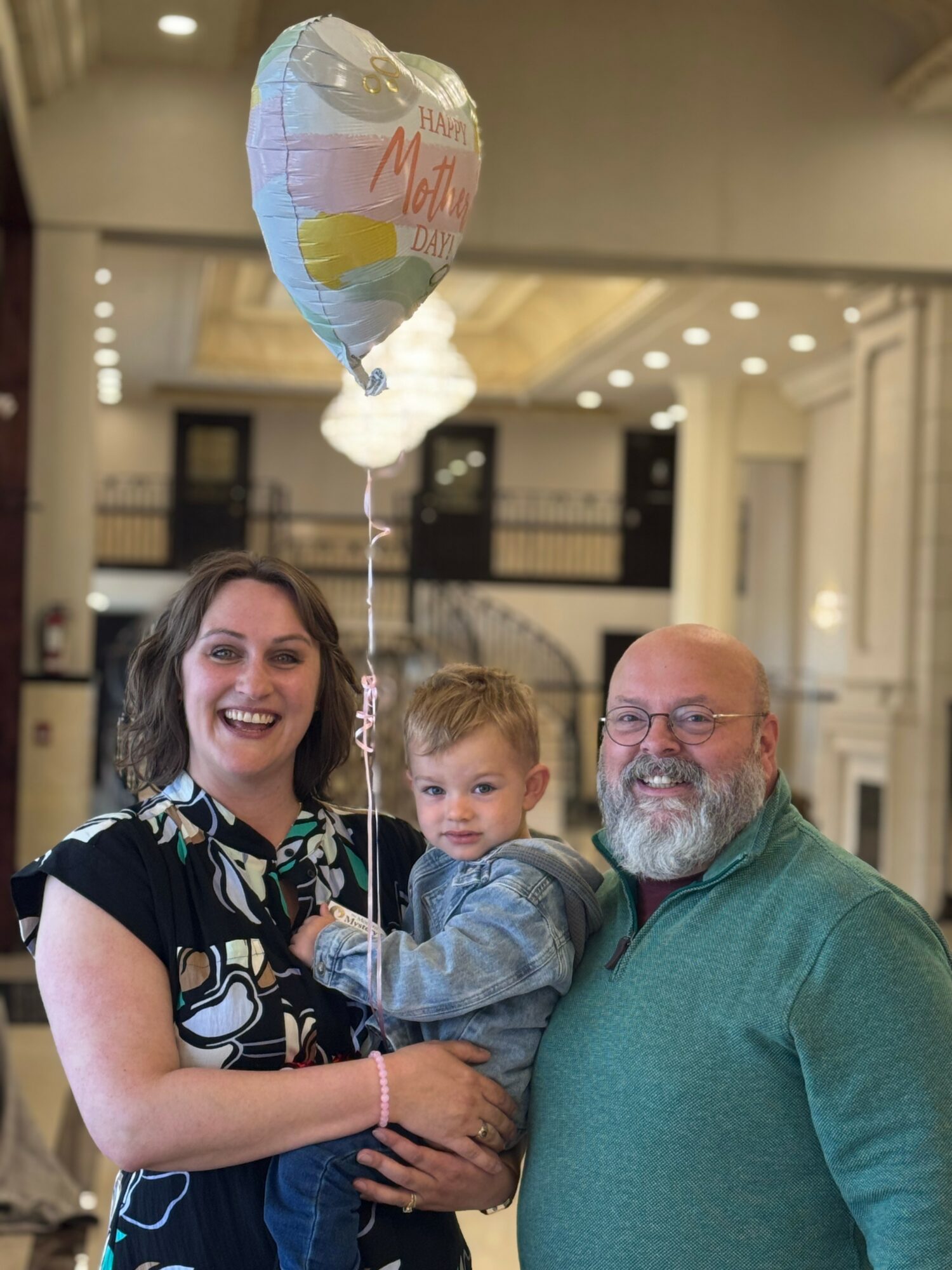 Three people, a woman, a man, and a child, smiling indoors with a balloon, in a spacious, well-lit area.