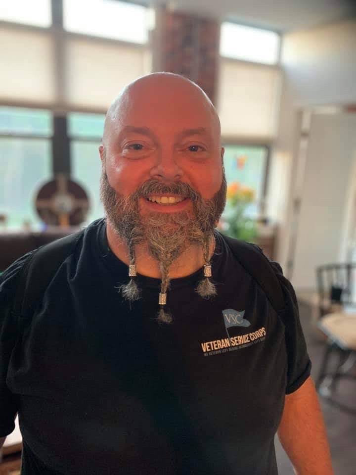 Bald man with beard smiling indoors, wearing a black shirt with a patch, in a room with large windows and furniture.