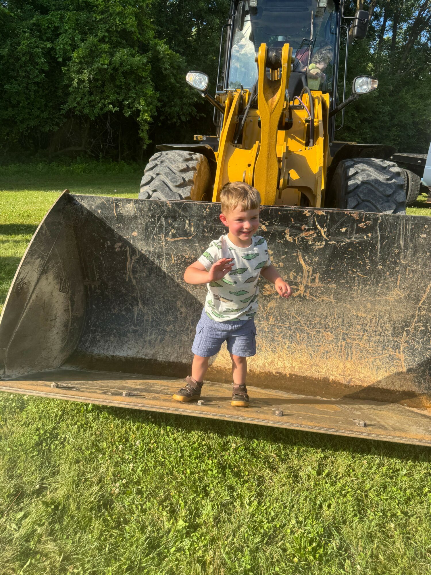 Child standing on a tractor bucket, holding a toy, outdoors with green grass and trees in background.