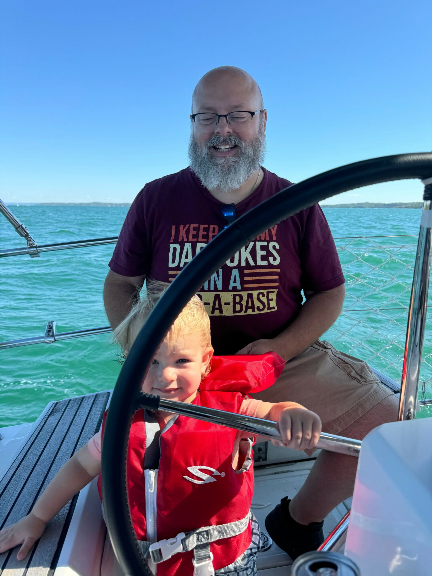 Two people on a boat with water and sky in the background, one with a beard and glasses, the other with a red life jacket.