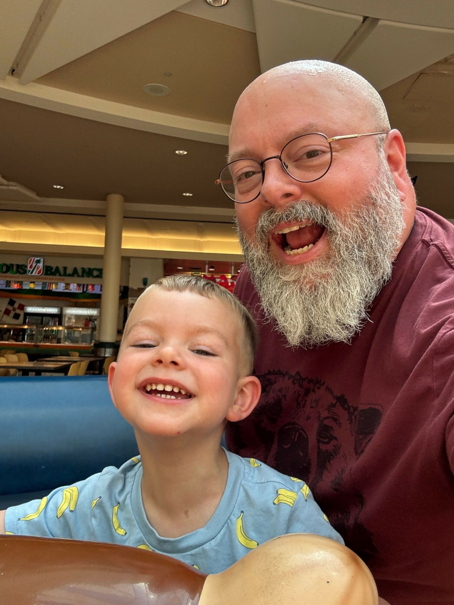 An older man with glasses and a beard and a young boy with a big smile sit together indoors.