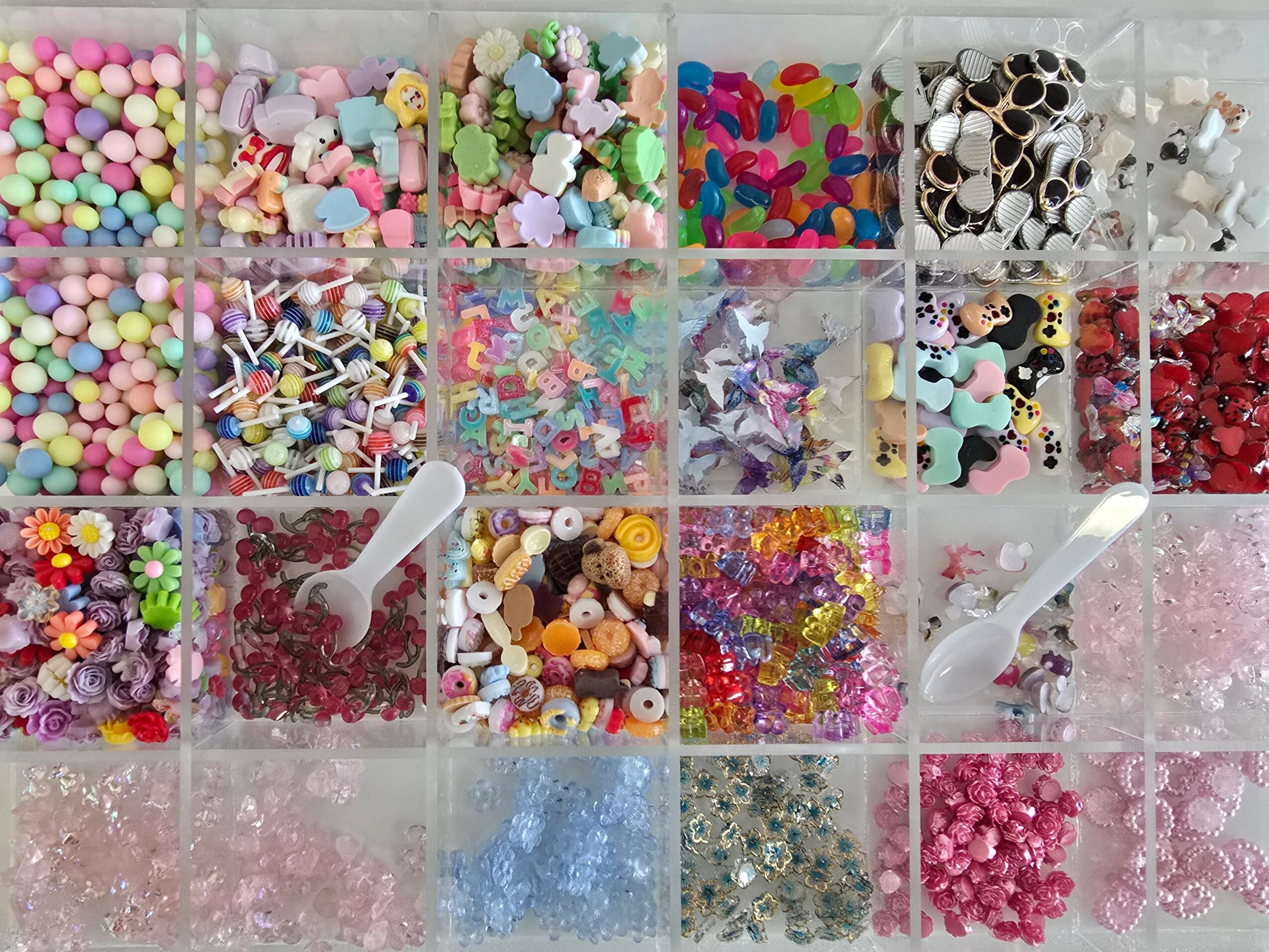 Multiple compartments filled with colorful craft supplies, including beads, sequins, and small decorative items, with two white scoops.
