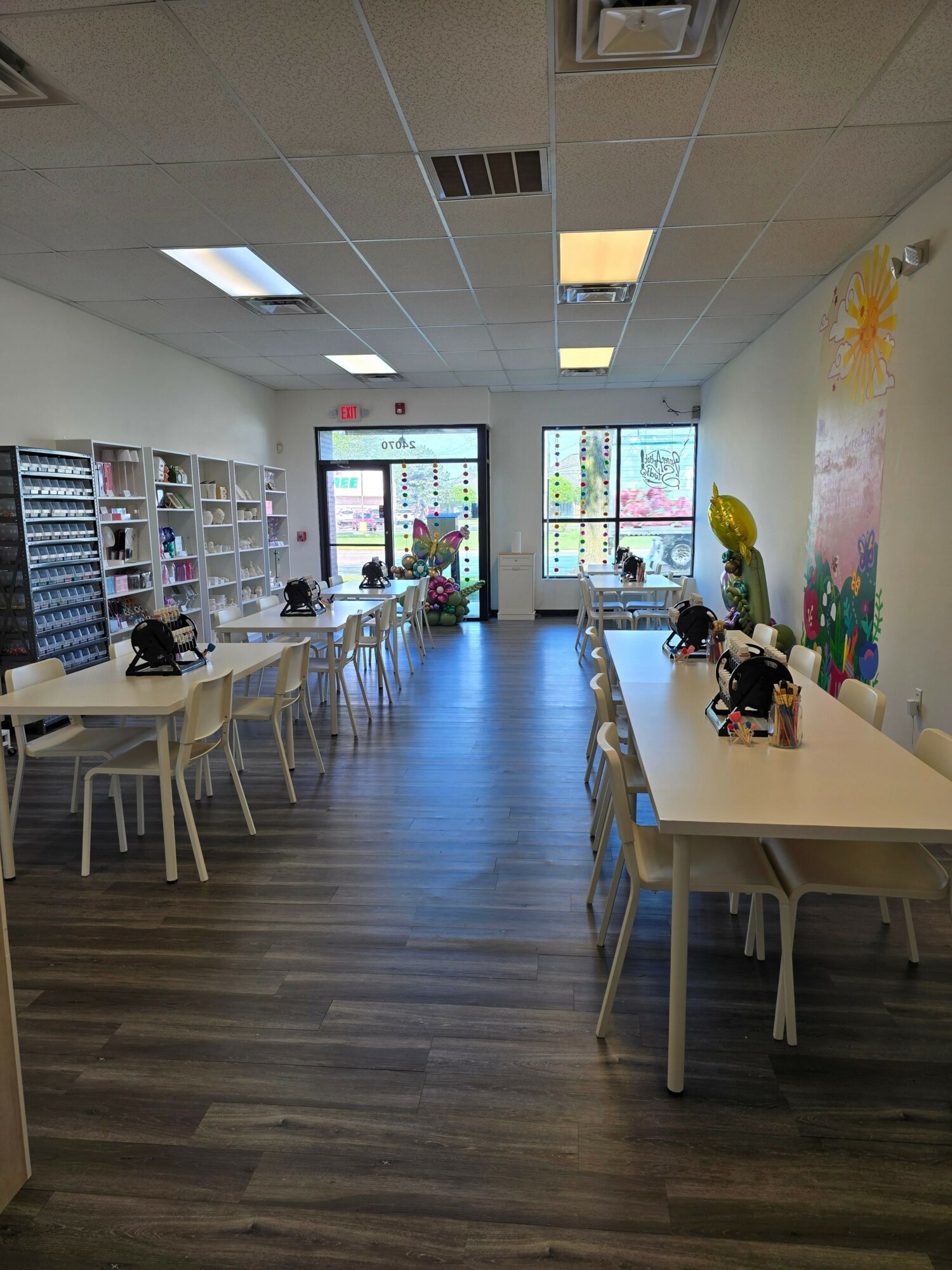 Room with white table, chairs, shelves, and large windows, decorated with plants and colorful artwork, wooden floor, ceiling lights.