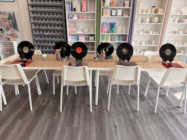 Table with five record players and chairs in a room with shelves of ceramics and supplies, gray flooring.