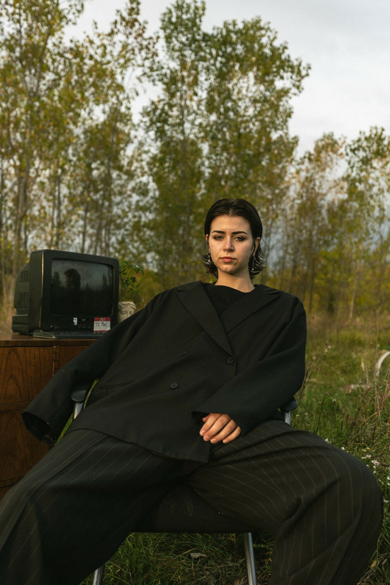 Woman in black oversized clothing sitting outdoors with trees and a black object in the background.