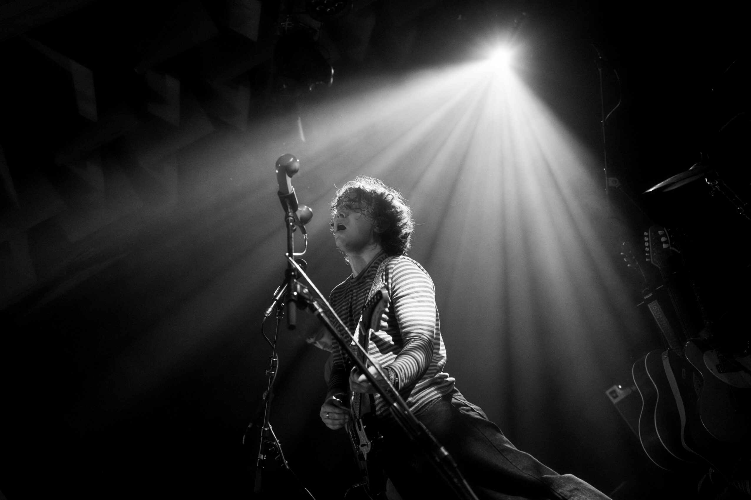 Person sitting on stage with a guitar, microphone, and stage lights shining down, in black and white.