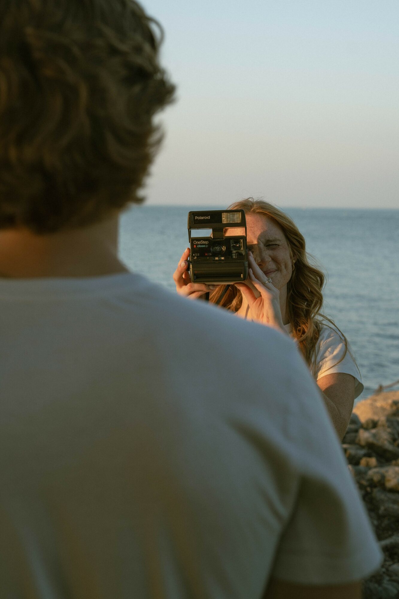 Person with curly hair holds a camera, capturing a woman by the water with a sunset sky in the background.