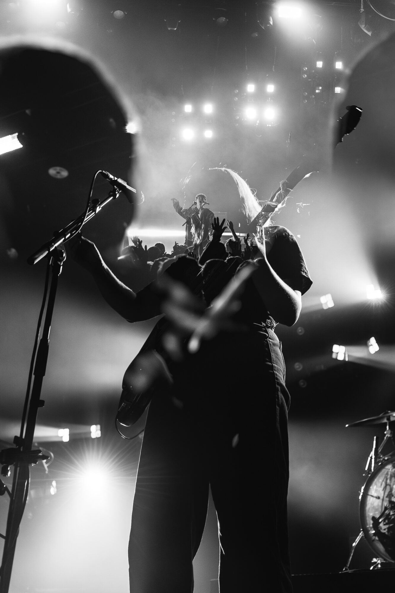 Musician on stage with microphone, playing guitar, illuminated by stage lights, with audience in background, black and white image.
