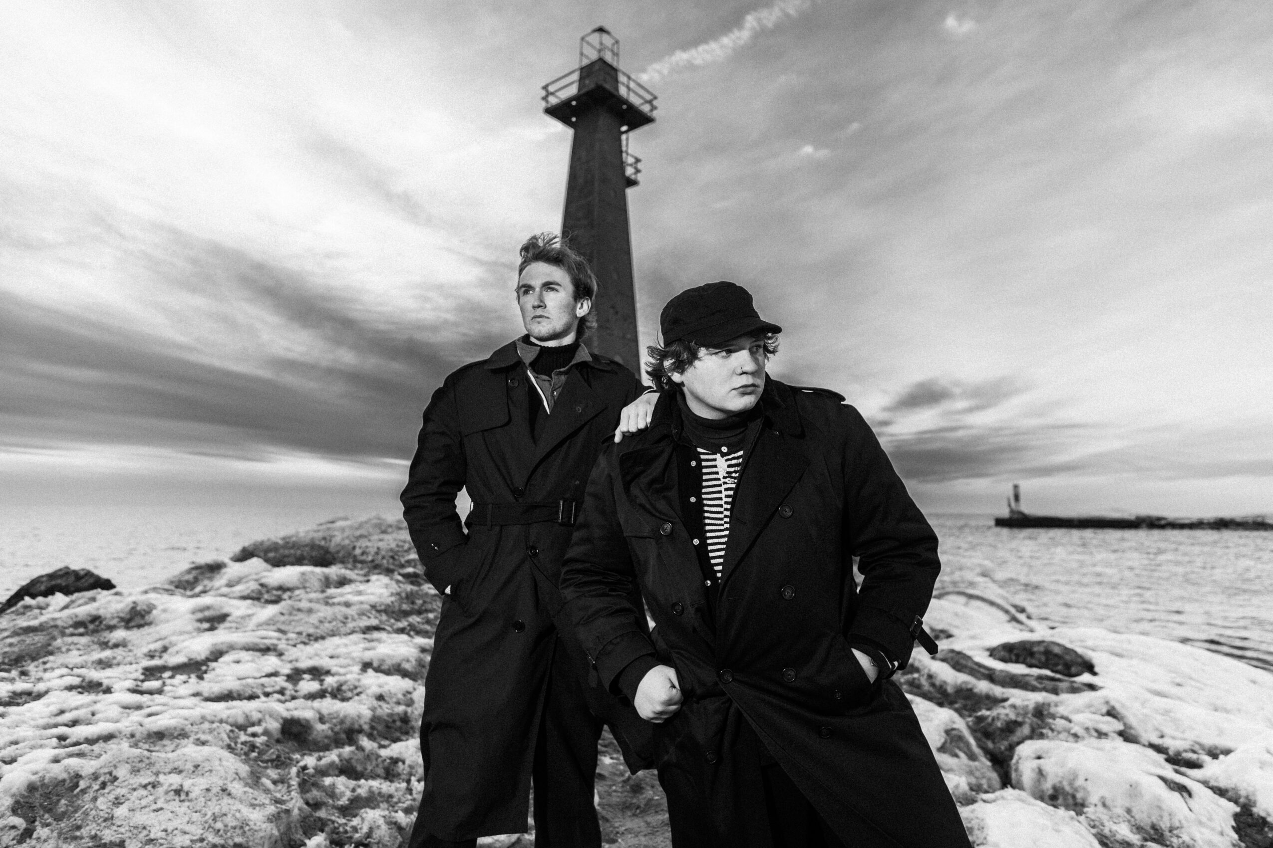Two people stand on rocks near the sea with a lighthouse in the background, cloudy sky above, in black and white.