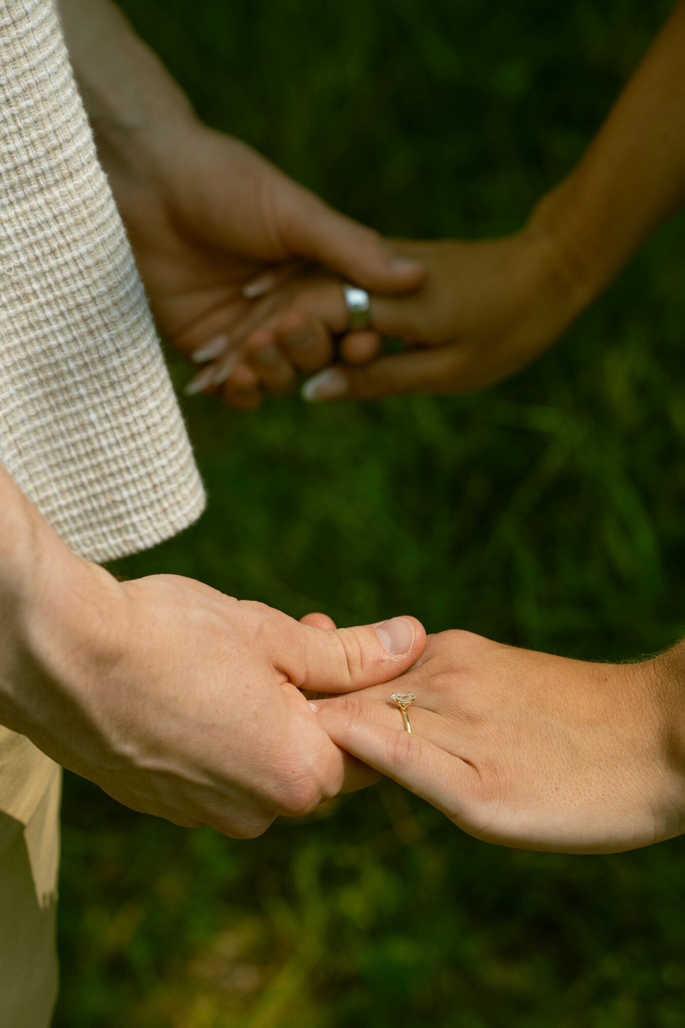Two pairs of hands holding each other, with a grassy background, one pair with a ring, in natural light.