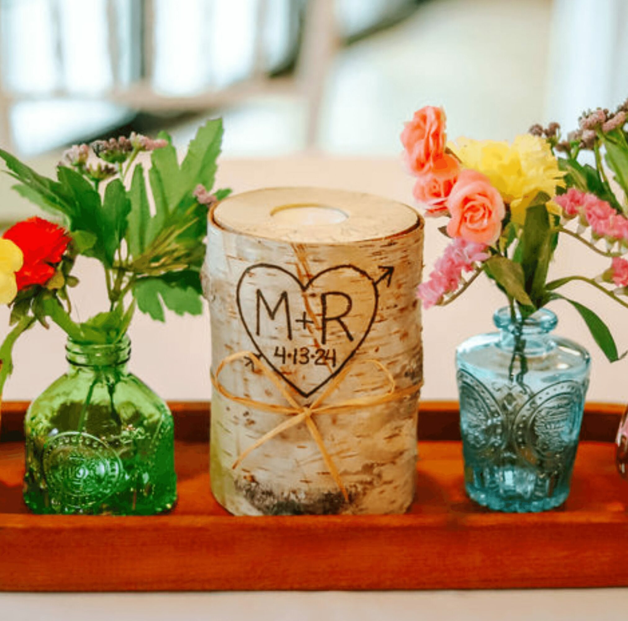 Decorative wooden candle holder with carved initials and date, flanked by colorful flower arrangements in glass bottles on a wooden tray.