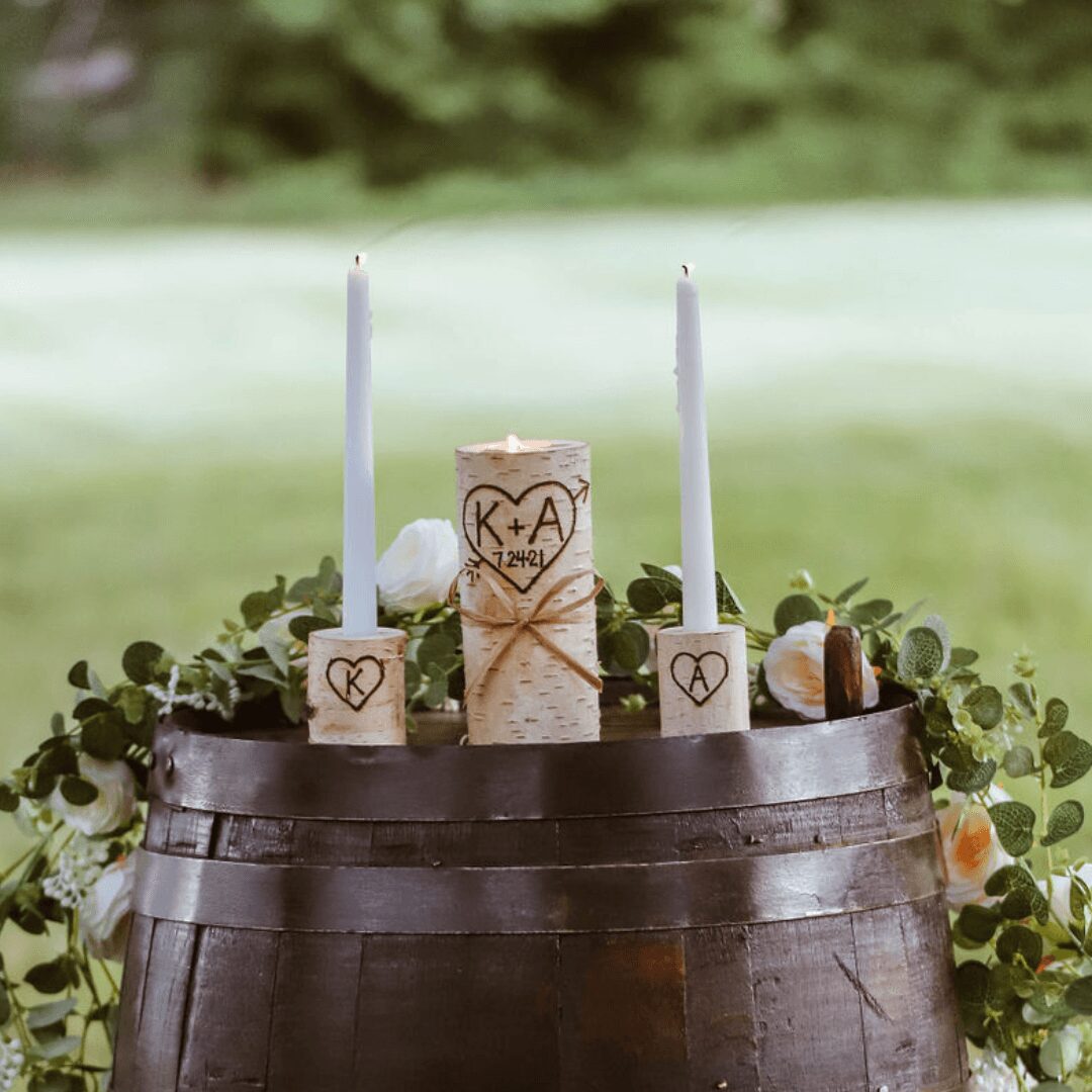 A wedding cake decorated with candles, surrounded by greenery and flowers, on a wooden barrel outdoors.