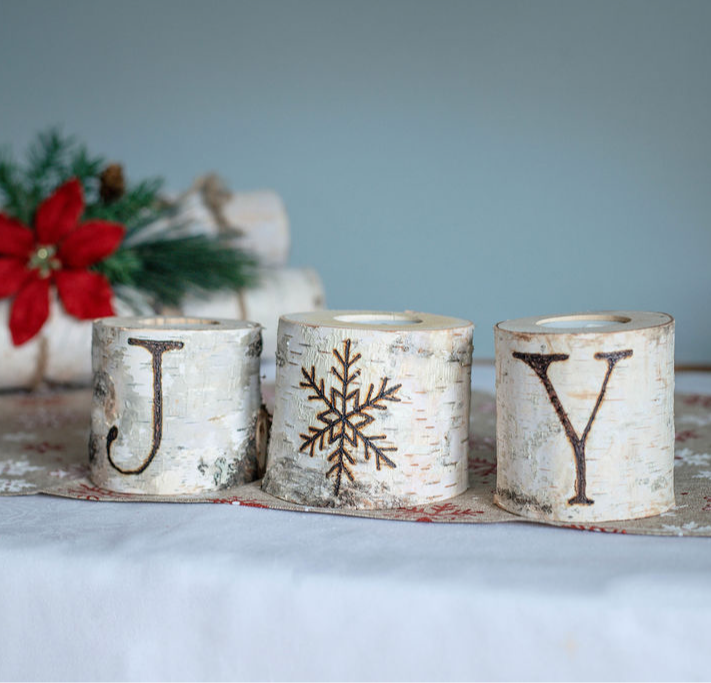 Wooden logs with painted letters J, snowflake, and Y, arranged on a table with holiday decorations in background.