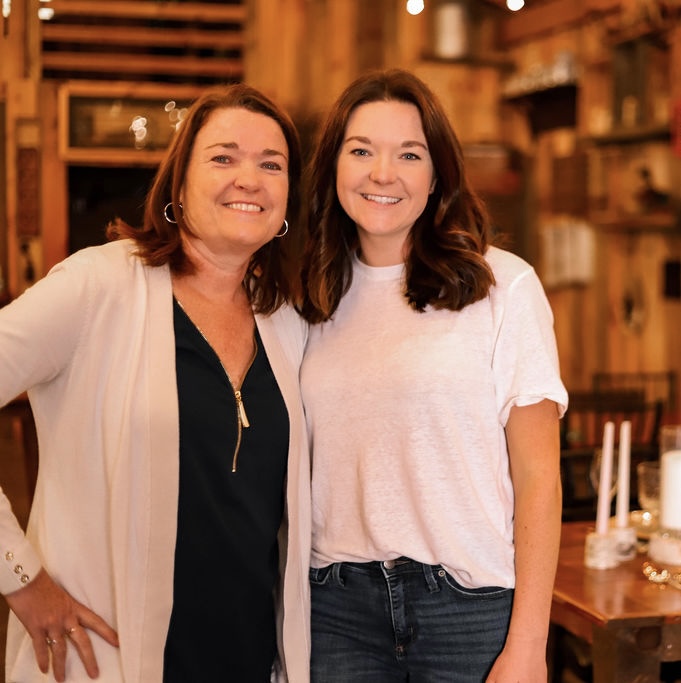 Two women smiling, standing close together indoors with wooden shelves and tables in the background.