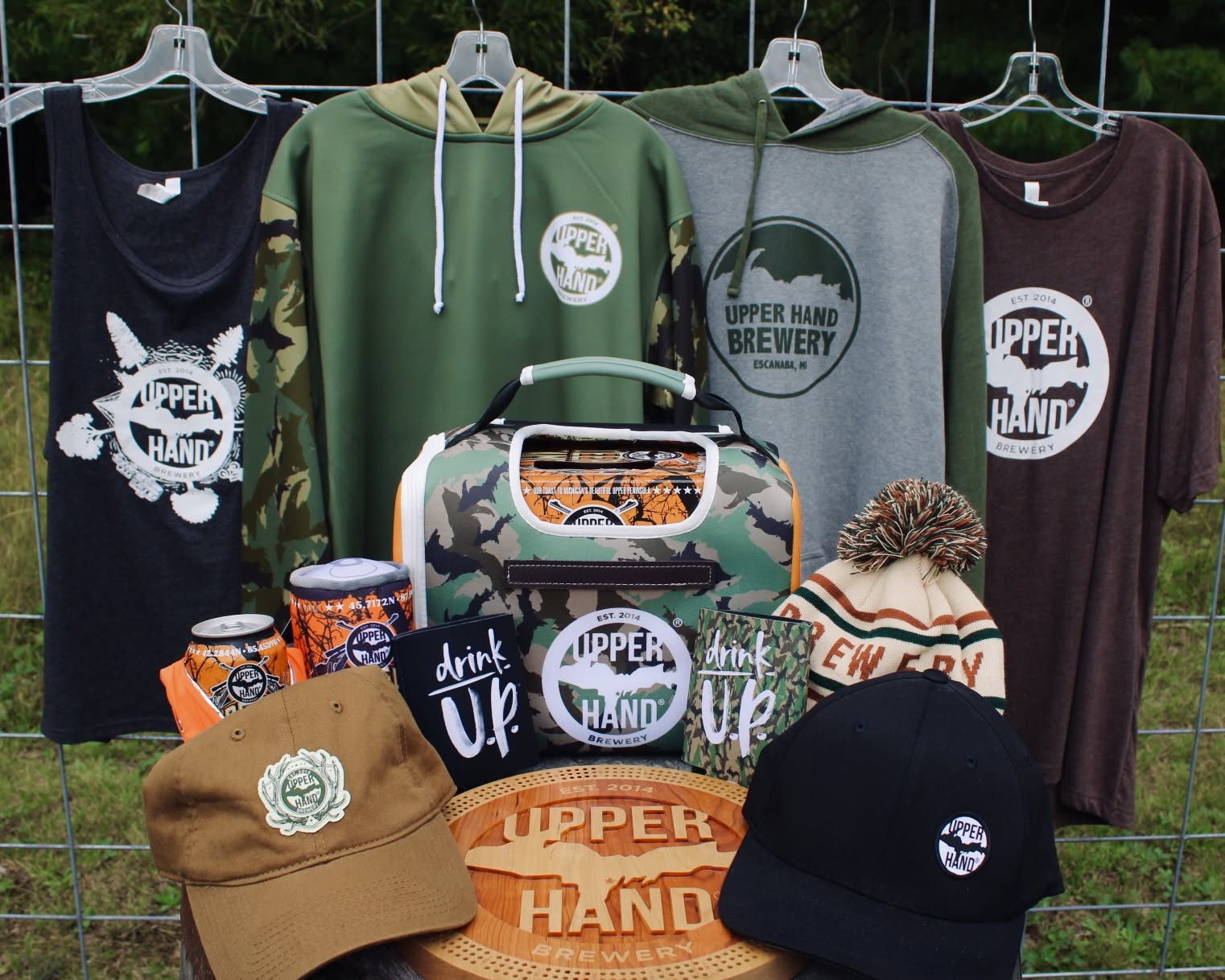 Clothing and accessories with brewery logos displayed on a wire rack outdoors, including hoodies, t-shirts, hats, and a bag.