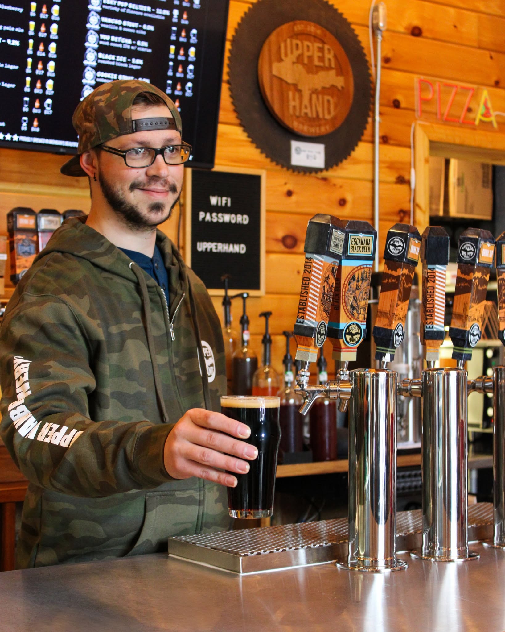 Man with glasses and a cap pours dark beer at a bar with taps, wooden wall, and a sign in the background.