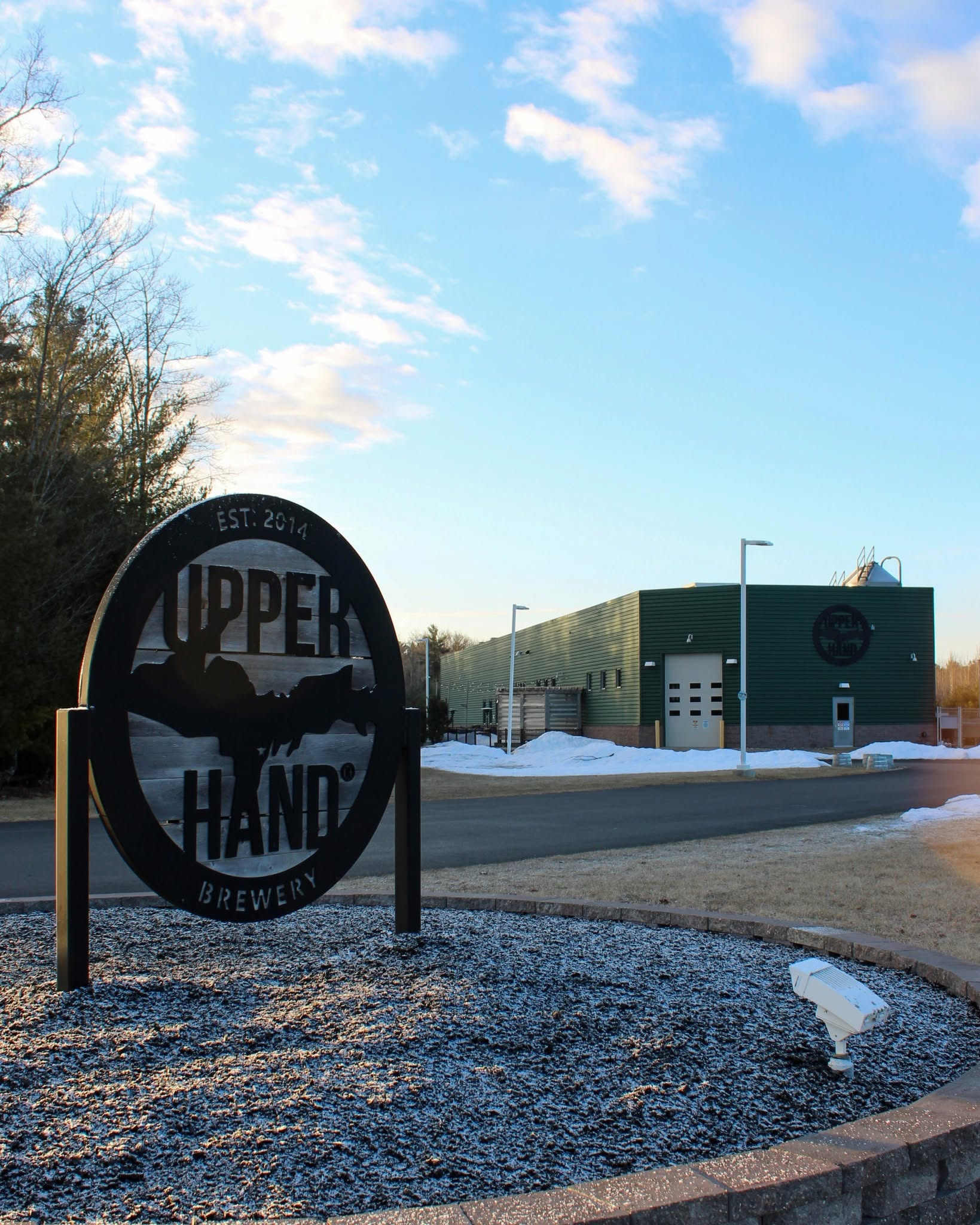 Outdoor scene with a sign reading 'Upper Hand Brewing' and a modern building in the background, snow on the ground, and a partly cloudy sky.