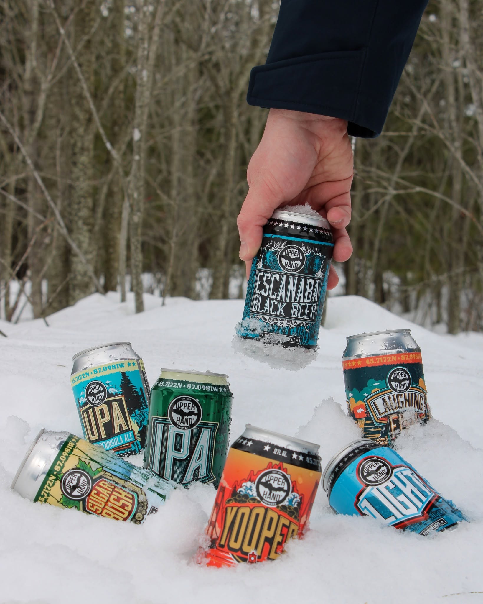 Person holding a can of Escanaba Black Beer outdoors in snow, with six other cans on snow, trees in background.