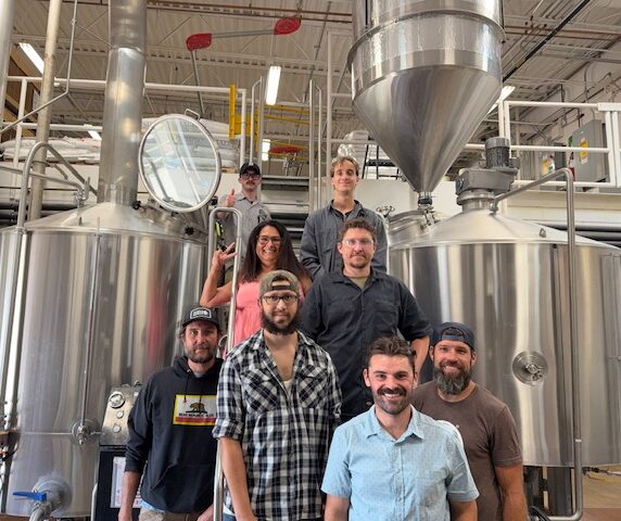 Group of eight people standing in front of large stainless steel industrial equipment in a factory or brewery setting.