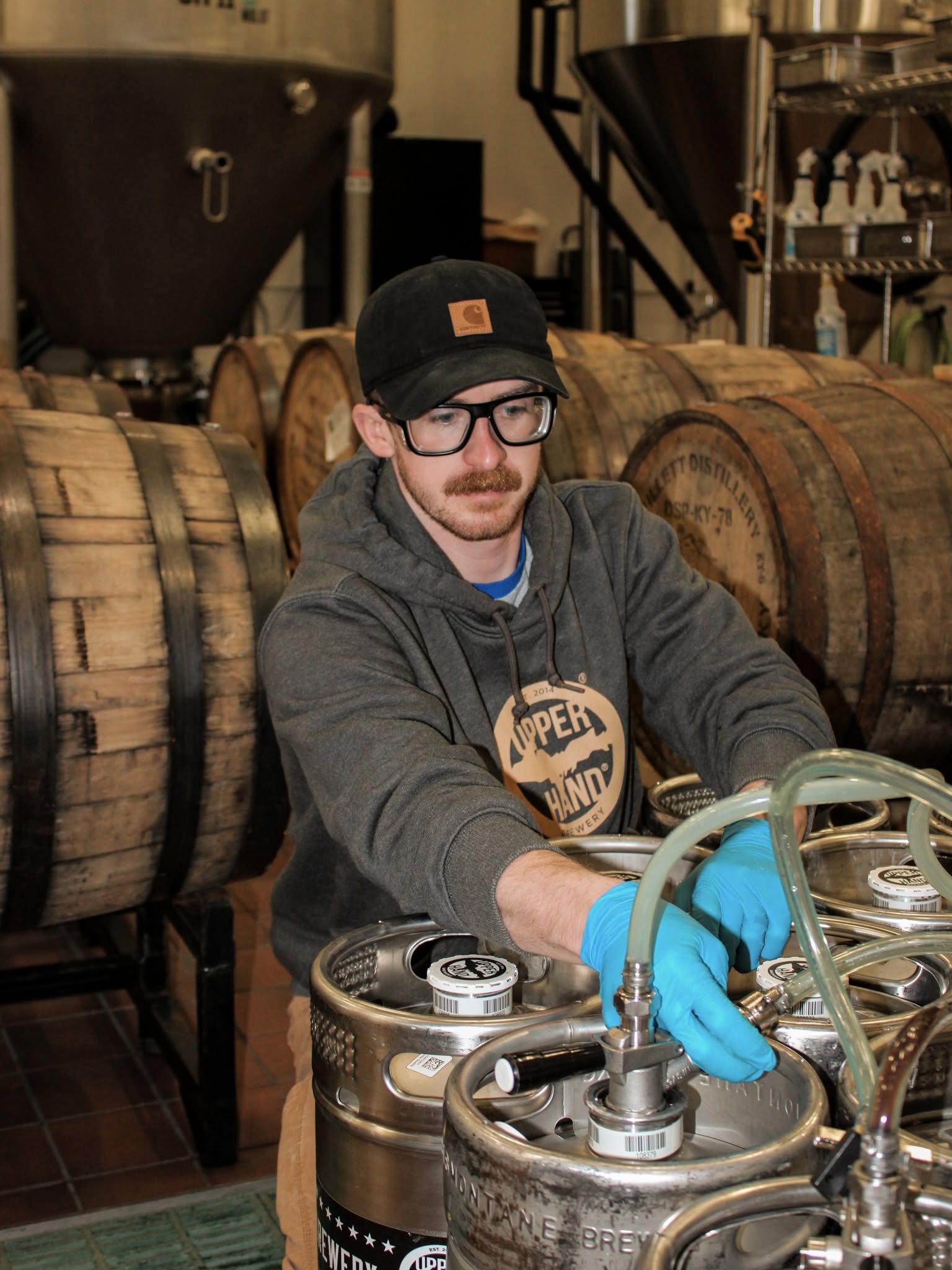 Man with glasses, black cap, and gray hoodie working with brewing equipment in a brewery, surrounded by wooden barrels.