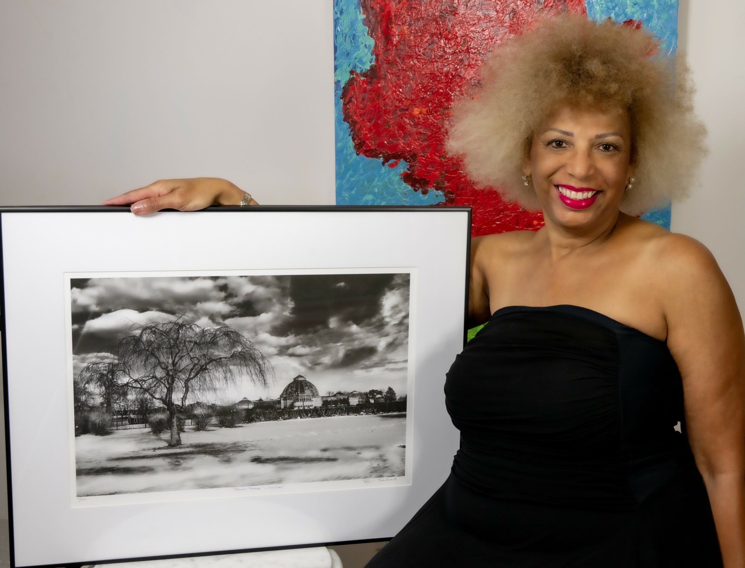 Woman with curly hair in a black strapless dress stands next to a framed black-and-white landscape photograph.