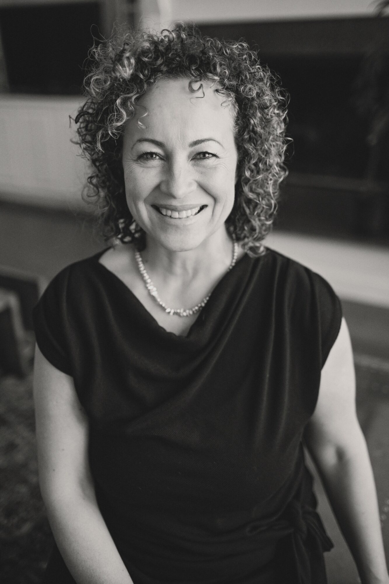 Smiling woman with curly hair wearing a dark top and necklace, indoors, in black and white.