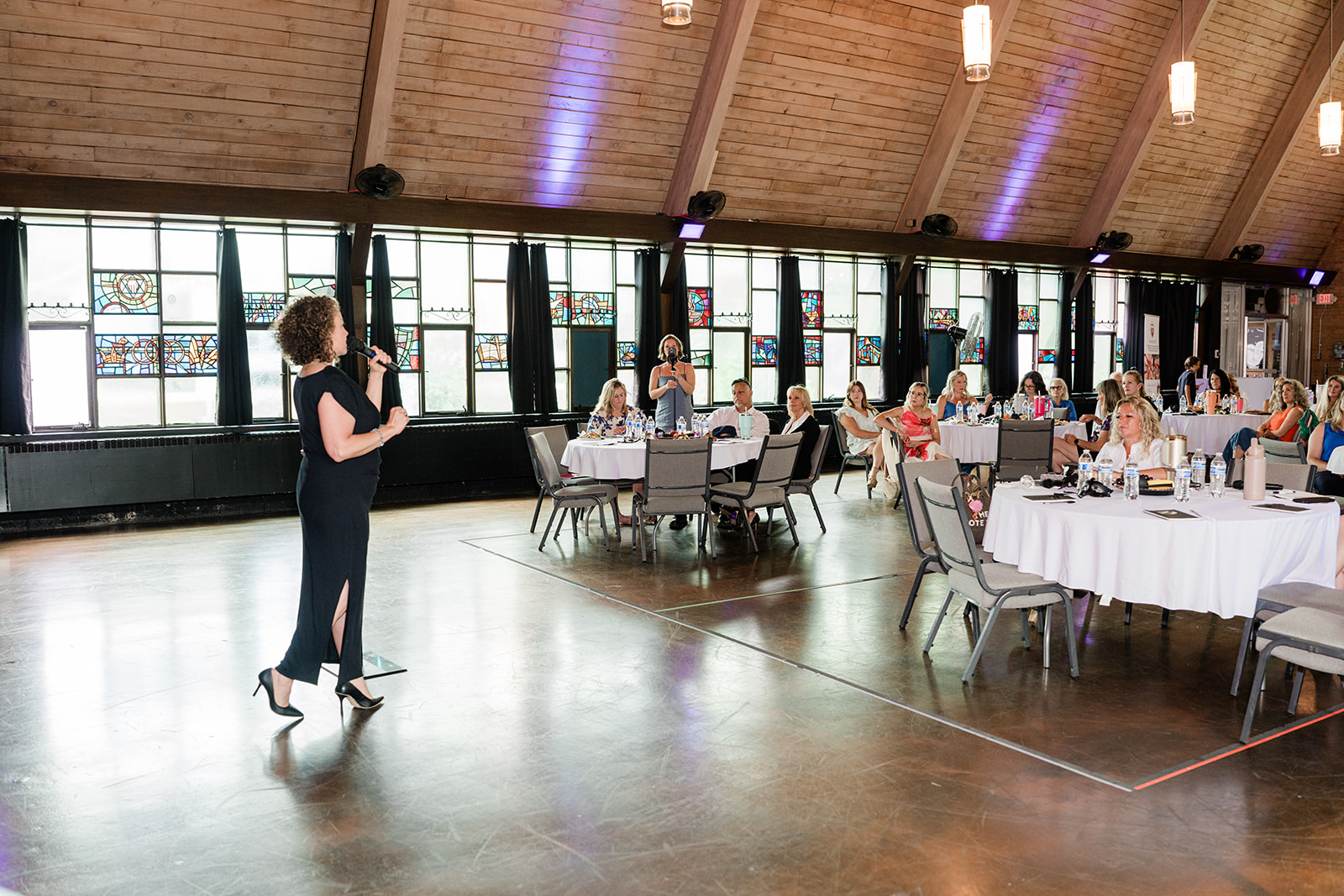A woman stands and speaks to a group of people seated at round tables in a spacious, well-lit event hall with large windows.